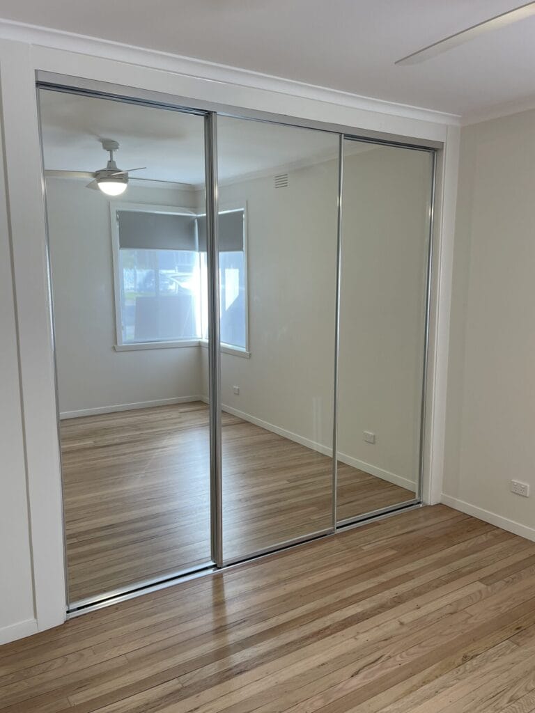 Sliding mirrored wardrobe doors in a bright room, reflecting a ceiling fan and window blinds across a light hardwood floor.