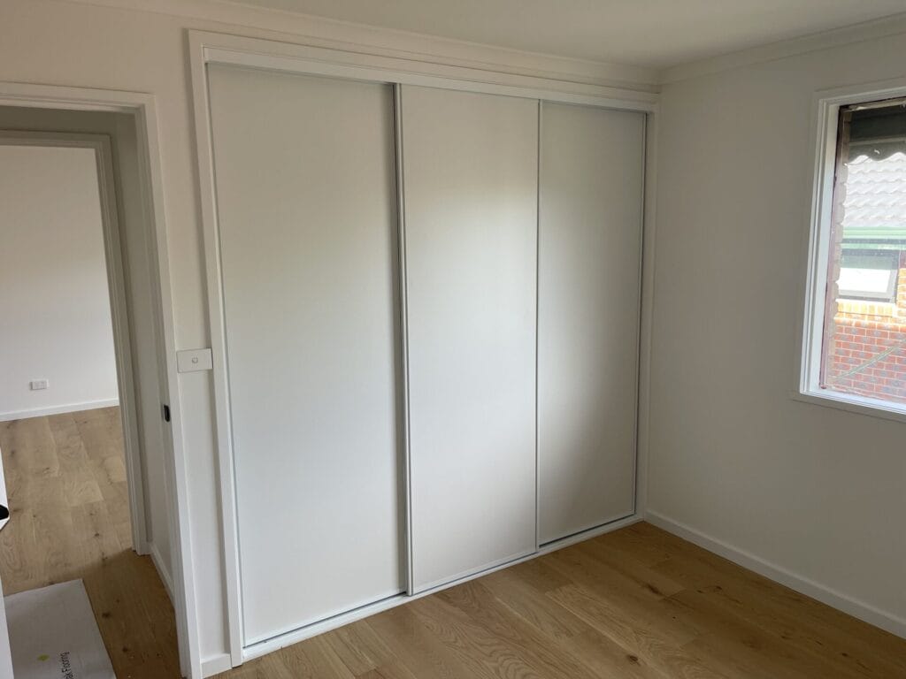 Three-panel white sliding-door wardrobe along a wall in a bright room, with a window to the right and wooden floor.